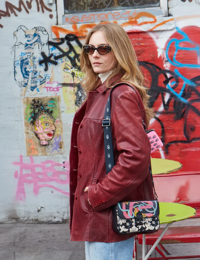 Woman in sunglasses and a red leather coat stands in front of a graffiti-covered wall, carrying a black shoulder bag with floral and serpent embroidery, and a pink pouch with embroidered eyes.