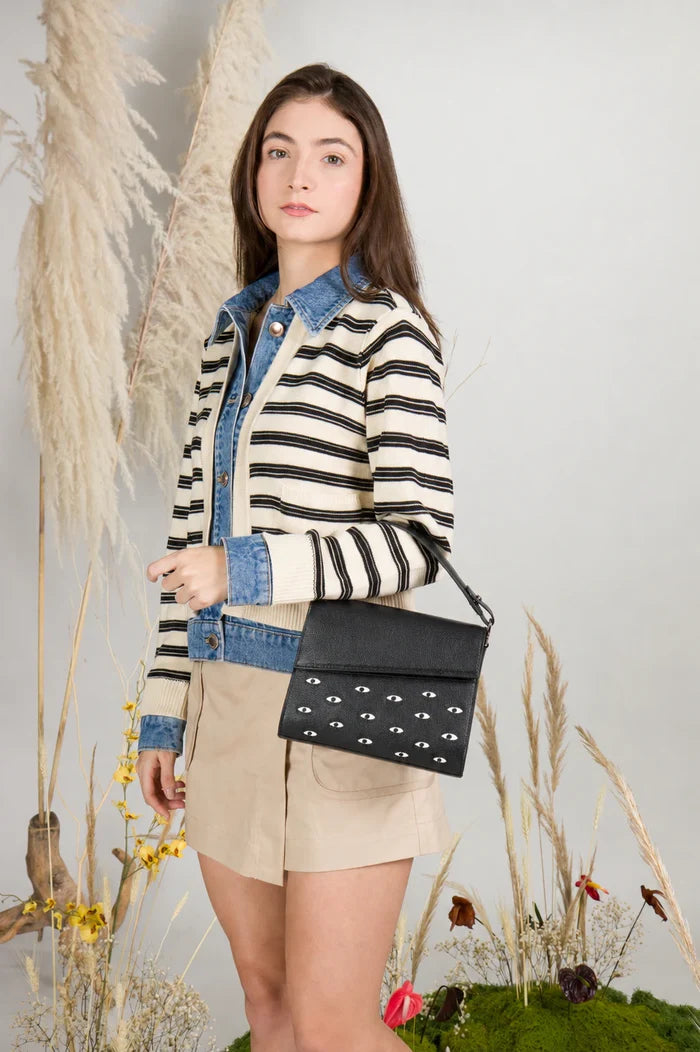 Young woman in a striped jacket and beige skirt poses in a botanical studio, wearing a black shoulder bag with white eye motifs.
