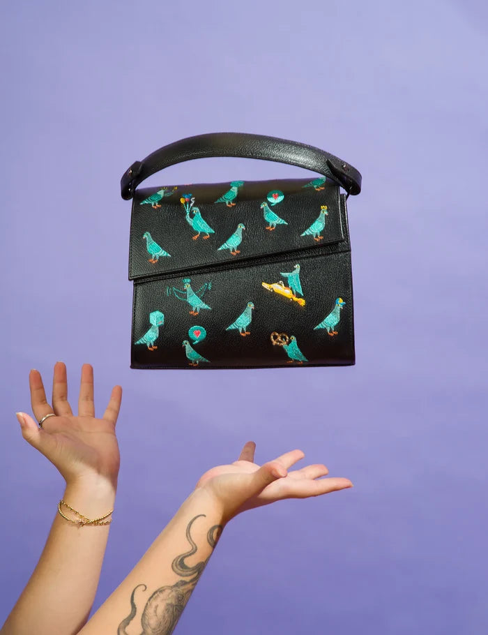 Tattooed arms toss a black handbag embroidered with playful teal pigeons, a pretzel, and a taxi, against a purple background. The bag floats mid-air with a curved top handle.