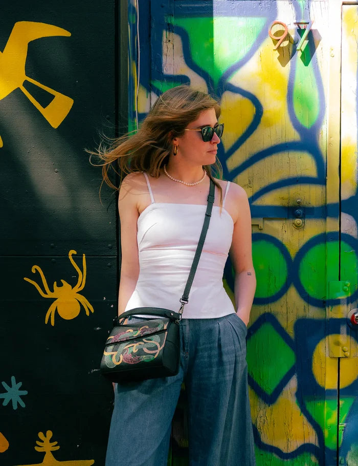 Woman in a white camisole and loose blue pants stands against a colorful graffiti wall, wearing sunglasses and a black crossbody bag with tigers and snake print design.