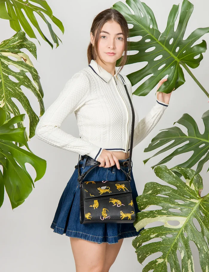 Woman in a white textured zip-up top and denim skirt poses among large green leaves, carrying a black handbag decorated with embroidered yellow tigers and pink snake details.
