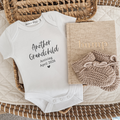 White baby onesie with text 'another grandchild' (script font), 'Arriving April 2026' in times roman. The onesie is laying in a woven basket with some beige baby booties and a linen beige bump journal.