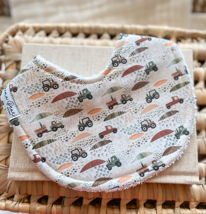 Curved handmade baby bib with a neutral farm print featuring green and brown tractors, red utes, and scattered fields in warm earth tones. The bib is backed with terry towelling and displayed on a beige cloth and woven basket.