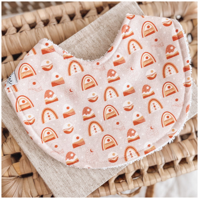 Handmade baby bib with a blush pink base and a playful pattern of terracotta-toned rainbows, suns, moons, and abstract shapes, backed with soft terry towelling.