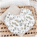 Handmade baby bib with a eucalyptus-themed print featuring green and grey leaves and sprigs on a white background. Bib is backed with beige terry towelling and styled on a woven basket.