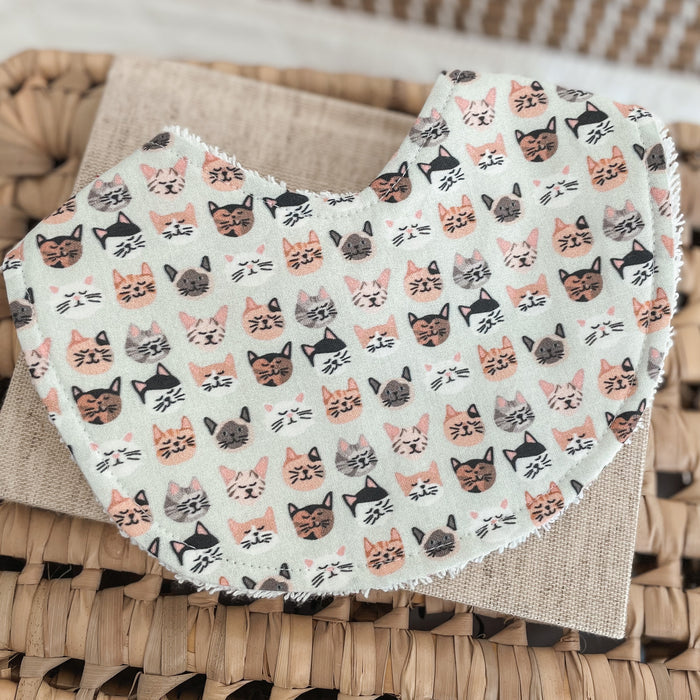 Flatlay of a handmade baby bib with a kitten face print. The design shows rows of illustrated cat faces in blush, grey, black, and peach shades on a light mint or soft sage background. Bib is backed with white terry towelling for added absorbency.