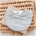 Seafoam blue baby bib featuring white curved line waves in a layered, ripple-style pattern, resting on neutral-toned fabric in a wicker basket.
