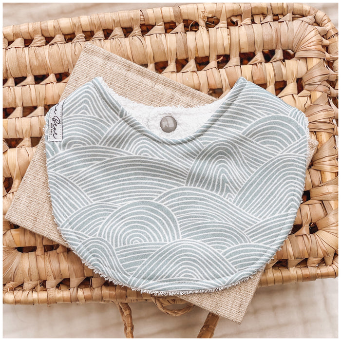 Seafoam blue baby bib featuring white curved line waves in a layered, ripple-style pattern, resting on neutral-toned fabric in a wicker basket.