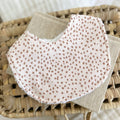 Neutral baby bib with terracotta fawn-like spots on a beige background. Handmade with cotton and terry towelling backing, displayed flatlay on woven mat.