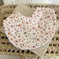Floral baby bib with blush, apricot, and beige flowers on a cream background, displayed on a neutral woven basket. Ideal for newborns, dribble, and teething.