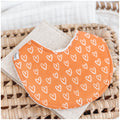 Handmade baby bib with a bright orange background and scattered white heart pattern. Bib is backed with beige terry towelling and displayed on a woven basket.