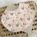 Flatlay of a handmade baby bib with a romantic floral pattern. The design includes pink and peach flowers with soft green leaves on a light cream base. Bib is made from cotton with an absorbent white terry towelling back.