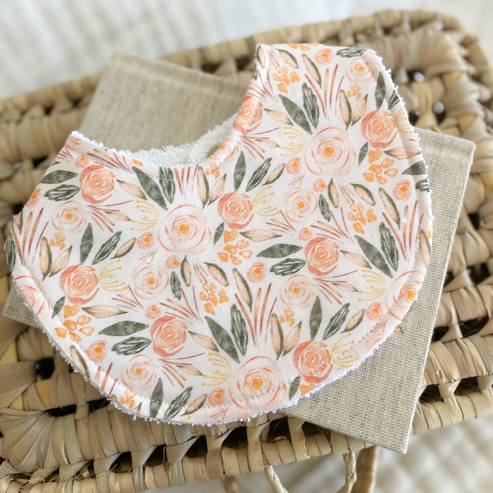 Flatlay of a handmade baby bib with a romantic floral pattern. The design includes pink and peach flowers with soft green leaves on a light cream base. Bib is made from cotton with an absorbent white terry towelling back.