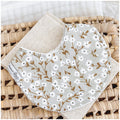 Baby bib featuring white dainty floral sprigs with golden brown stems on a muted sage grey background, displayed on a linen cloth and wicker basket.
