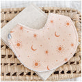 Handmade baby bib with a soft peach background and scattered burnt orange sun, moon, and star illustrations. Bib is backed with beige terry towelling and styled on a woven basket.