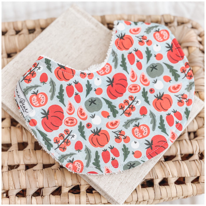 Handmade baby bib with a colourful vegetable print featuring tomatoes, rocket leaves, and sliced produce on a soft pale blue background.