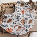 Baby bib with autumn woodland print featuring owls, moths, leaves, and berries in warm earthy tones, laid on a woven basket background.
