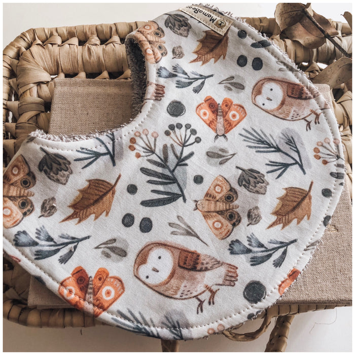 Baby bib with autumn woodland print featuring owls, moths, leaves, and berries in warm earthy tones, laid on a woven basket background.