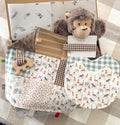 A gift box full with baby gifts including a brown monkey toy comforter, a dark brown check gingham dummy clip, a blue gingham bib, illustrations of jungle animals on another bib and a patchwork taggie blankie with wooden elephant teether. The taggie blankie has mainly light brown, mustard, blue tones with a chocolate brown soft mini fabric on the back. Some of the animals shown in the fabrics are: zebra, giraffe, lion, elephant, monkey, cheetah.