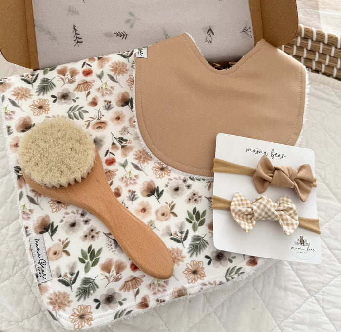 Baby girl gift box including a beige bib, a floral burpcloth, a wooden baby brush, and two bow headbands.
