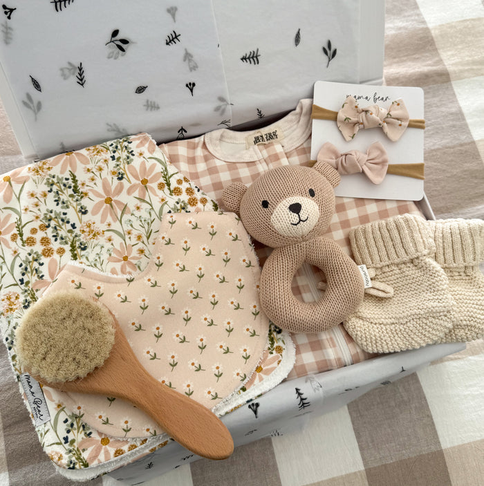 Luxury baby girl gift box featuring a soft pink gingham zip growsuit, handmade floral baby bib, matching floral burp cloth, two blush baby headbands, knitted teddy bear baby rattle, cream knitted baby booties and a natural wooden baby hair brush, beautifully arranged inside a gift box with white botanical tissue paper. Perfect as a newborn baby girl gift or baby shower present.
