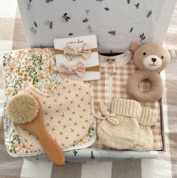 Beautiful newborn baby girl hamper displayed in a keepsake gift box with blush pink and neutral baby essentials including a pink gingham zip growsuit, floral handmade bib and burp cloth set, knitted teddy bear rattle, cream baby booties, wooden baby brush and matching baby headbands. A thoughtful baby shower gift, newborn gift or pregnancy gift for a baby girl.