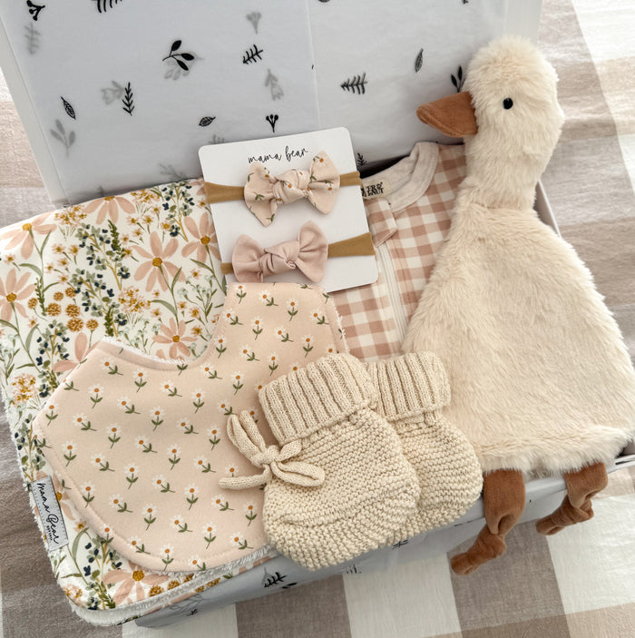 Luxury baby girl gift box featuring a soft pink gingham zip growsuit, handmade floral baby bib, matching floral burp cloth, two blush baby headbands, cream knitted baby booties and a soft plush goose comforter, beautifully arranged inside a keepsake gift box with white botanical tissue paper. A thoughtful newborn baby girl gift, baby shower present or maternity leave gift.
