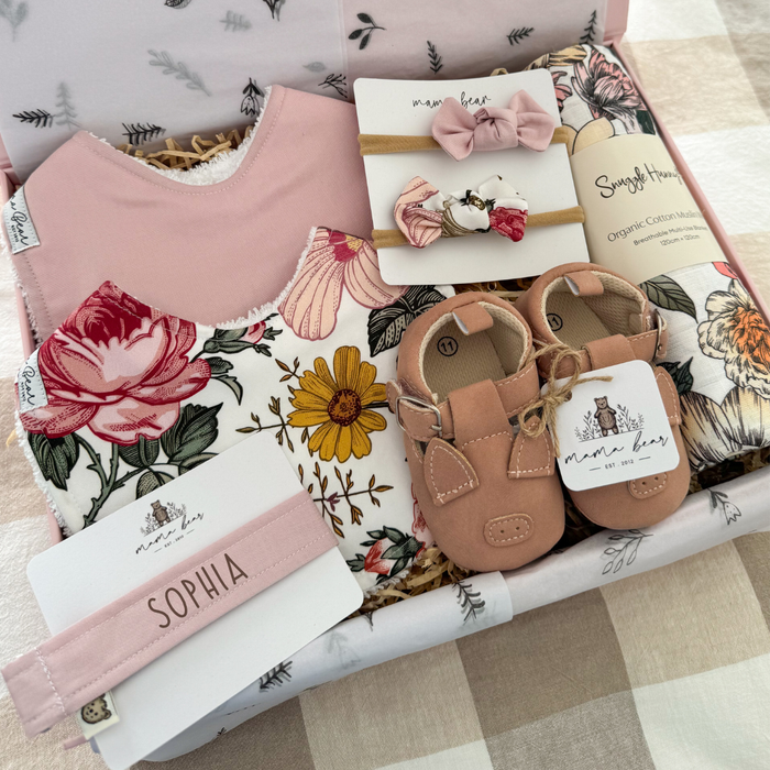 Pink Baby girl gift hamper featuring a solid pale pink bib, another bib with scattered roses and daisy flowers in pinks and mustards, two matching hairbows, a pair of baby shoes with pink piggy faces and a floral swaddle  in pink, pale yellow and green tones.