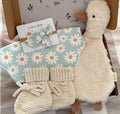 A baby gift set arranged in a kraft box, featuring a plush cream goose toy, a aqua and white daisy-patterned bib with scalloped edges, a matching daisy-patterned baby bow and a pair of cream knit booties.