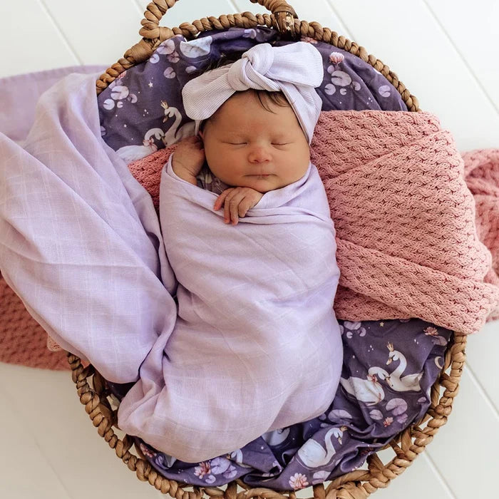 Newborn baby swaddled in a purple blanket with a floral headband, lying in a woven basket with pink and purple blankets.