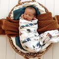 Newborn baby swaddled in a blanket with train pattern, wrapped in brown fabric, lying in a woven basket.