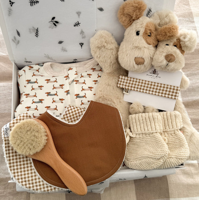 Neutral newborn baby gift box featuring a dachshund print zip growsuit, handmade baby bibs in warm brown gingham & a solid tan shade, natural wooden baby hair brush, cream knitted baby booties, soft plush cream dog comforter, matching puppy rattle. Beautifully presented in a keepsake gift box, perfect for a baby shower or newborn gift.