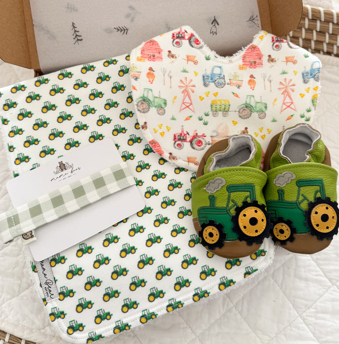 A baby gift set featuring a green tractor patterned burp cloth, a matching bib, a green and white gingham dummy clip, and a pair of green tractor theme baby shoes.
