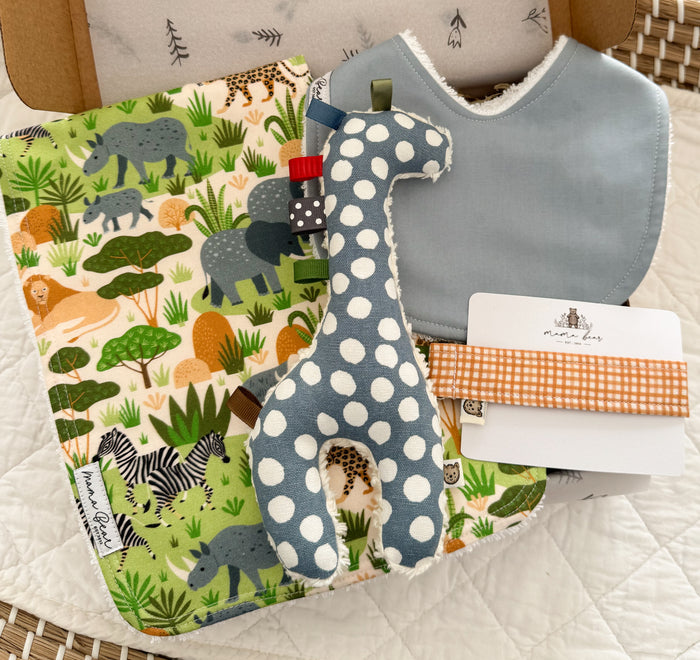 A baby gift box featuring a vibrant jungle theme burp cloth, a light blue fabric bib, a blue and white spot giraffe toy, and an orange gingham dummy clip.