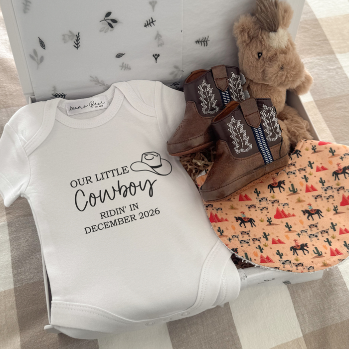cowboy pregnancy announcement gift box featuring a white “Our Little Cowboy Ridin’ In December 2026” baby onesie, plush horse rattle, brown baby cowboy boots and a handmade cowboy print baby bib with horses, cacti and desert illustrations. A unique baby shower gift or pregnancy announcement gift for expecting parents.