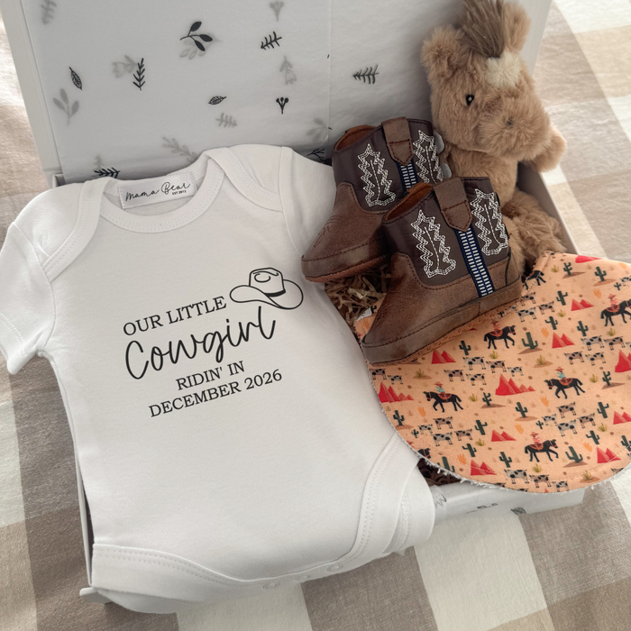Western-themed pregnancy announcement gift box featuring a white “Our Little Cowgirl Ridin’ In December 2026” baby onesie, plush horse comforter, brown baby cowboy boots and a handmade cowboy print bib with horses, cacti and desert illustrations. A unique baby shower gift or pregnancy announcement gift for expecting parents.