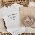Pregnancy announcement white baby onesie saying 'Grandma's Bestie' in black script font with a small heart. Linen Journal and beige baby booties as props in the flay lay photo.