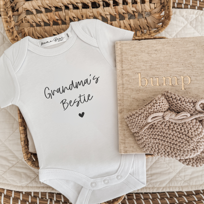 Pregnancy announcement white baby onesie saying 'Grandma's Bestie' in black script font with a small heart. Linen Journal and beige baby booties as props in the flay lay photo.