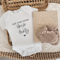 Flat lay of a white baby onesie printed with “See you soon Uncle + Aunty” in black script and serif fonts, with a small heart underneath. The onesie is styled in a wicker basket with a beige “bump” pregnancy journal and a pair of soft knitted baby booties. A customisable baby announcement onesie, perfect for sharing the news with loved ones like grandparents, aunties, uncles, or personalised names.