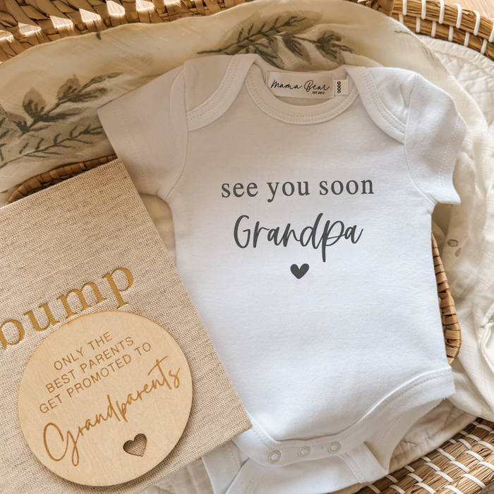 Flat lay of a customisable white baby onesie printed with the phrase “See you soon Grandpa” in mixed serif and script fonts with a heart beneath. The onesie is styled in a wicker basket with a leafy muslin wrap, a “bump” pregnancy journal, and a round wooden plaque that reads “Only the best parents get promoted to grandparents.” A heartfelt pregnancy announcement onesie, perfect for surprising Grandpa—or any loved one—with customisable name options.