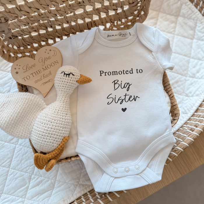 White baby onesie with 'Promoted to Big Sister' text, crocheted duck toy, and wooden heart ornament on a textured surface.