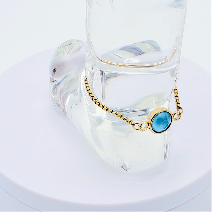 Gold penis bracelet with a blue gemstone on a clear glass stand against a white background