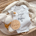 Flat lay of a personalised white baby onesie printed with “Can’t wait to meet you Yiayia and Pappou” in a mix of serif and script fonts with a small heart underneath. The onesie is styled inside a wicker basket on a leafy muslin wrap, paired with a white knitted goose toy and a round wooden plaque that reads “We’re having a baby.” A beautiful and customisable pregnancy announcement idea for announcing to grandparents, including in-laws or family members with cultural names.