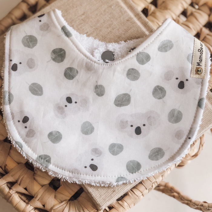 Handmade baby bib with soft grey koalas and green eucalyptus leaves on a white cotton background, backed with white terry towelling.