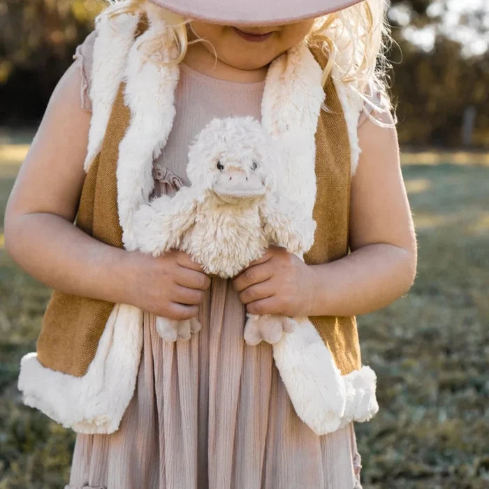 Child wearing a pink hat, brown vest, and beige dress holding a white duck in a natural setting.