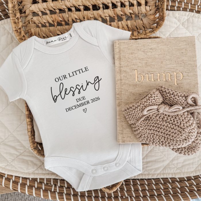 Australia baby announcement. White onesie with black vinyl text 'Our little blessing due December 2026' with a small little heart underneath. Photo is a flaylay with onesie pictured in a basket, with a beige linen pregnancy journal and beige knit booties.