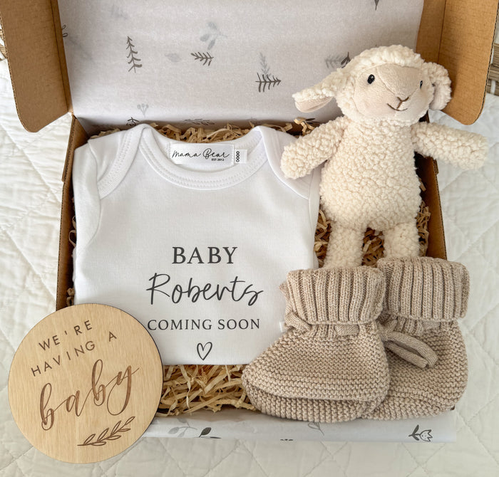 Pregnancy announcement gift idea for family. Personalised white baby onesie 'Baby Roberts Coming Soon' with a small heart. Baby lamb white toy rattle. Beige booties and a wooden plaque 'We're having a baby'.
