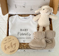 Pregnancy announcement gift idea for family. Personalised white baby onesie 'Baby Roberts Coming Soon' with a small heart. Baby lamb white toy rattle. Beige booties and a wooden plaque 'We're having a baby'.