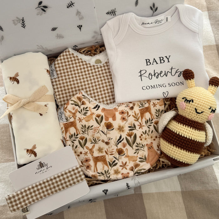 Baby gift box filled with beige gingham and woodland tones. Featuring a soft white swaddle with small illustrations of bumble bees, a beige and cream mini gingham bib with matching dummy clip, a bib with illustrations of woodland creatures and flora such as deer, bunny, bear, birds all in soft browns and sage.  A white baby onesie with black text personalised with 'BABY Robert coming soon' and a small heart underneath. And a crochet little bee baby comforter in a pale yellow and brown stripes wit white wing