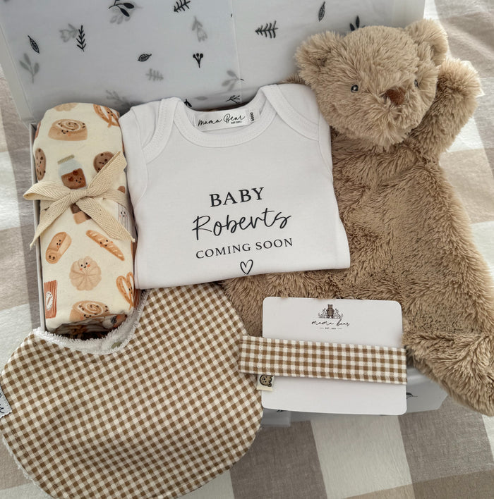 Baby shower gift set with personalsied baby onesie 'Baby Roberts coming soon', soft brown teddy bear comforter, brown gingham check baby bib & matching dummy clip and a cream swaddle with cute illustrations of bakery foods such as danishes and bread. Packaged inside a white gift box.